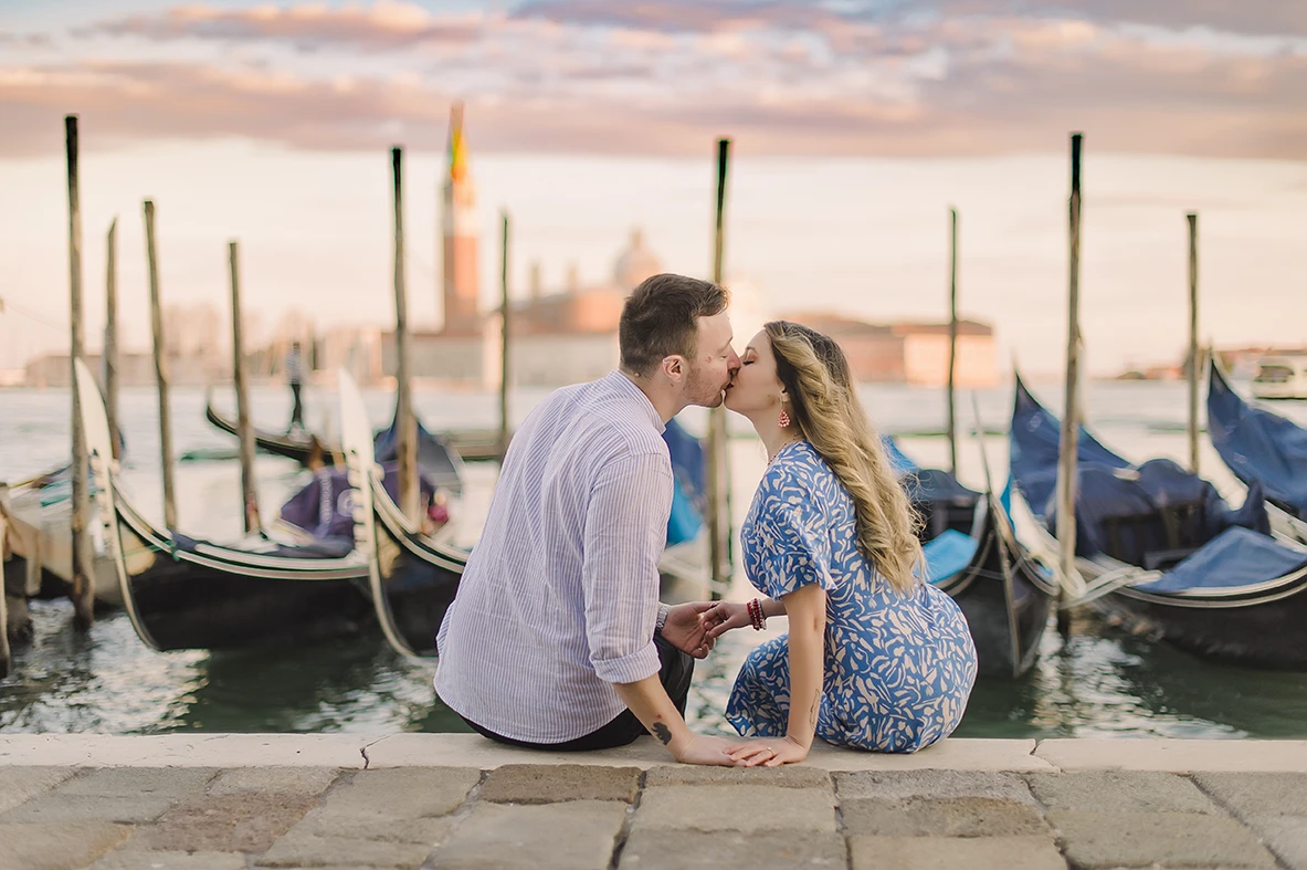 he fiancés share a gentle kiss  with the historic buildings of Venice and boats passing by in the soft, fading light of dusk