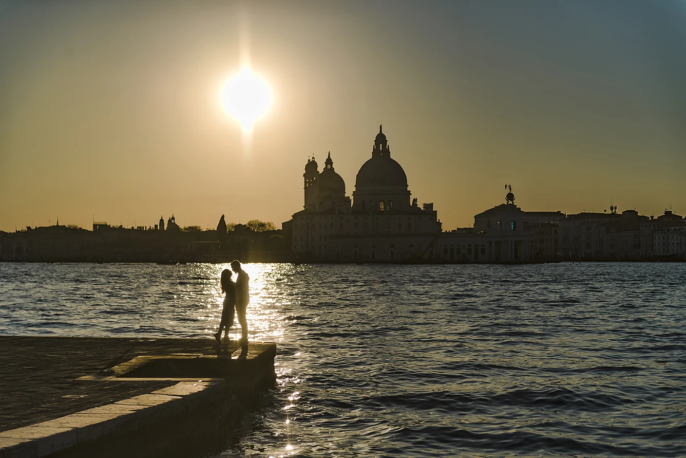 An intimate kiss of Barth and Maria as they stand by the Grand Canal, their silhouettes framed against the twinkling lights of evening Venice