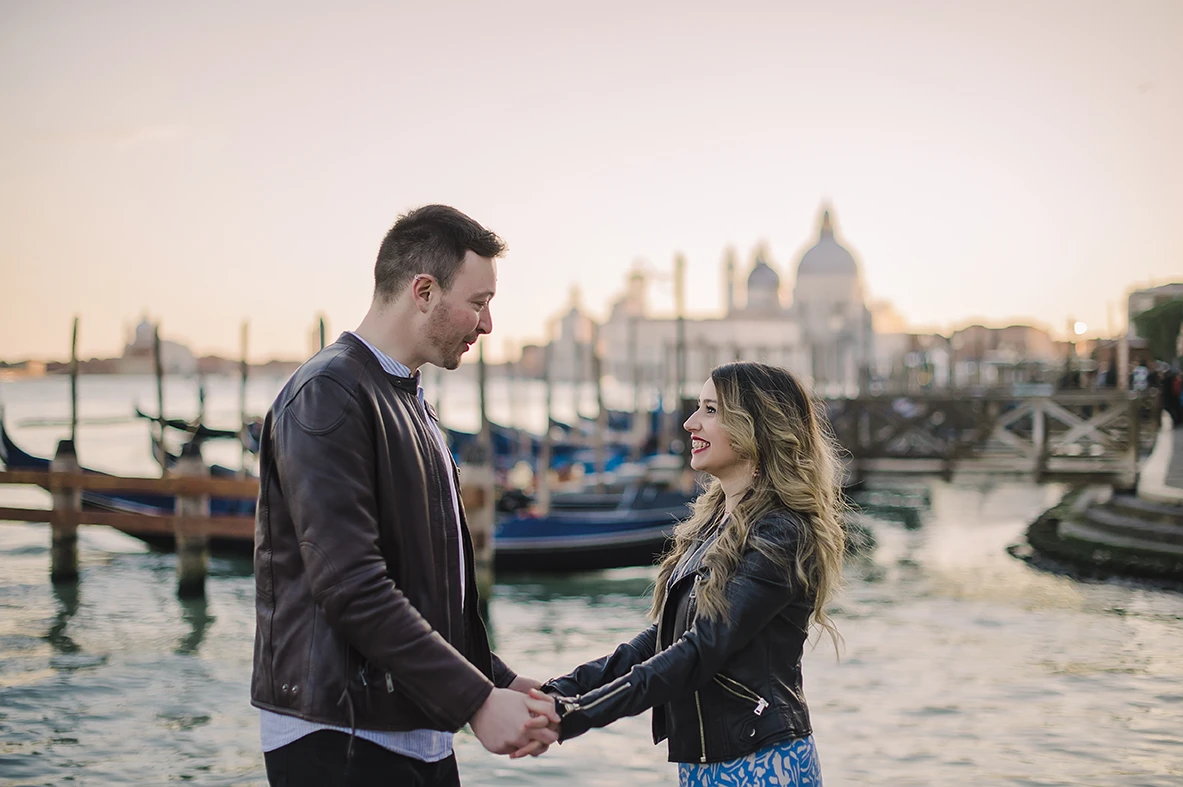 A candid shot capturing the couple's spontaneous dance, with Maria's flowing dress and the serene, amber-tinted sky of Venice creating a timeless scene.