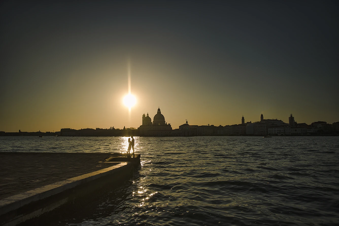 An intimate kiss of Barth and Maria as they stand by the Grand Canal, their silhouettes framed against the twinkling lights of evening Venice
