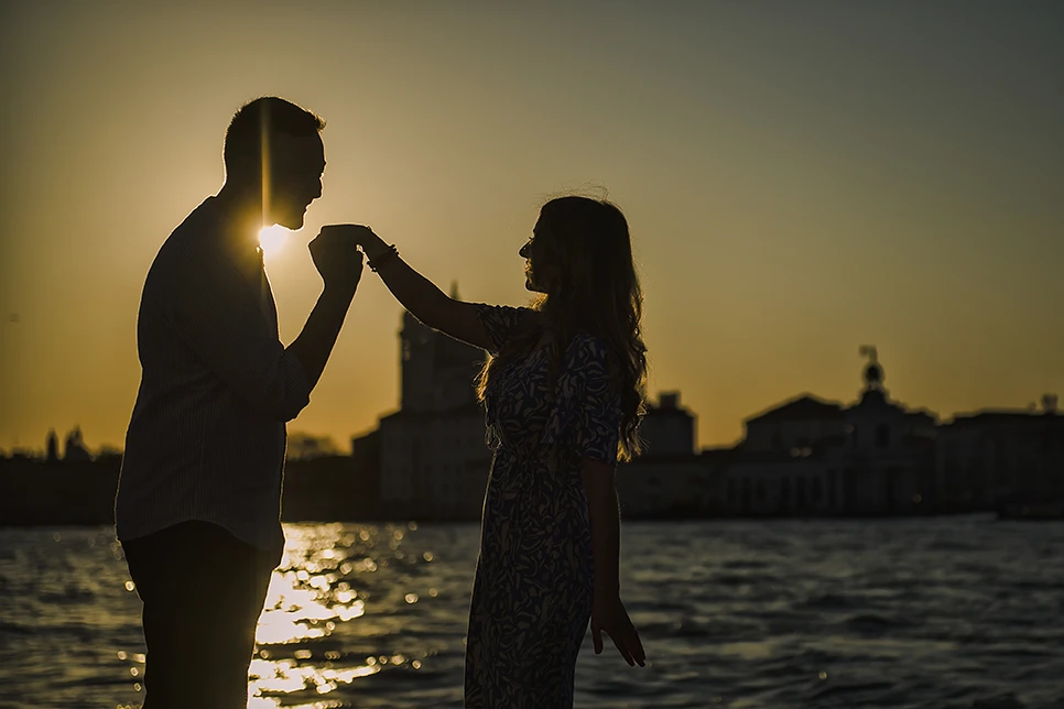 An intimate kiss of Barth and Maria as they stand by the Grand Canal, their silhouettes framed against the twinkling lights of evening Venice