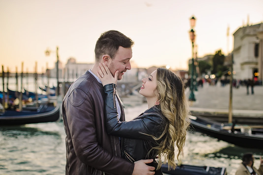 Laughing together, Maria playfully places her engagement ring-adorned hand on Barth's chest as they savor the moment, a gondola leisurely drifting in the background.