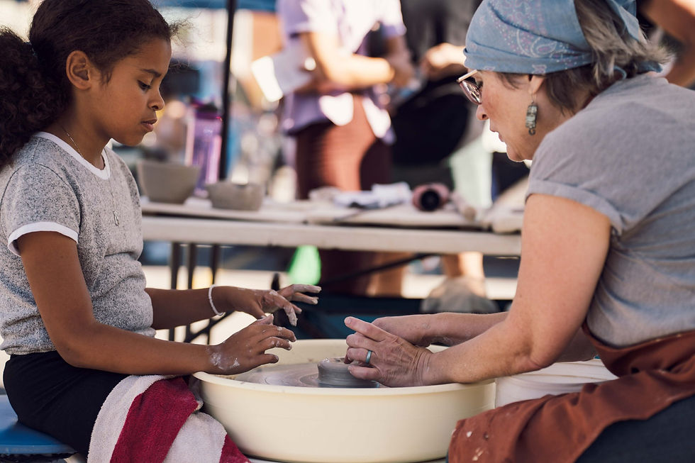 A woman shows a younger girl how to make a bowl out of clay on a pottery wheel outdoors.