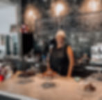 Owner Barbara stands behind the restaurant counter. Behind her is a chalkboard menu and in front of her are plates piled with different desserts.