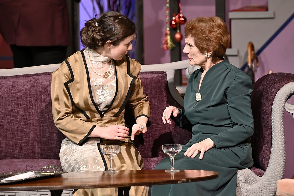 A young woman and older woman dressed in 1930s clothing talk on a couch.