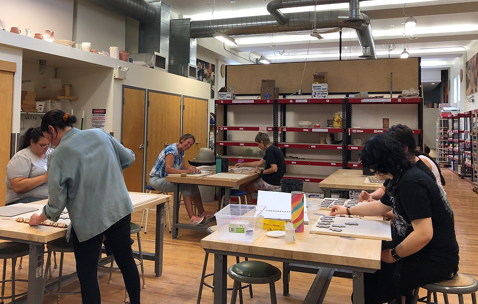 Several people scattered throughout a clay workshop working at tables. Some are standing, some are sitting.