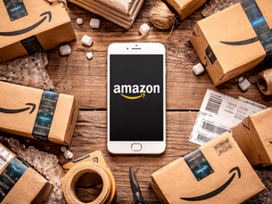 Top-down photograph of a white smartphone displaying the Amazon logo on its screen, placed on a rustic wooden table surrounded by Amazon shipping boxes, packing tape, labels, bubble wrap, and scissors, forming a realistic e-commerce and home delivery still-life scene