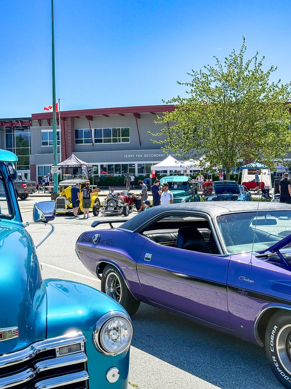 Classic cars, including a blue truck and purple Challenger, parked at Terry Fox Secondary School. People gather around under a clear sky.