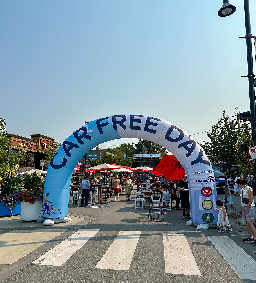 Outdoor event with people under a large "Car Free Day" arch. Red umbrellas and stalls in the background. Clear blue sky. Lively atmosphere.