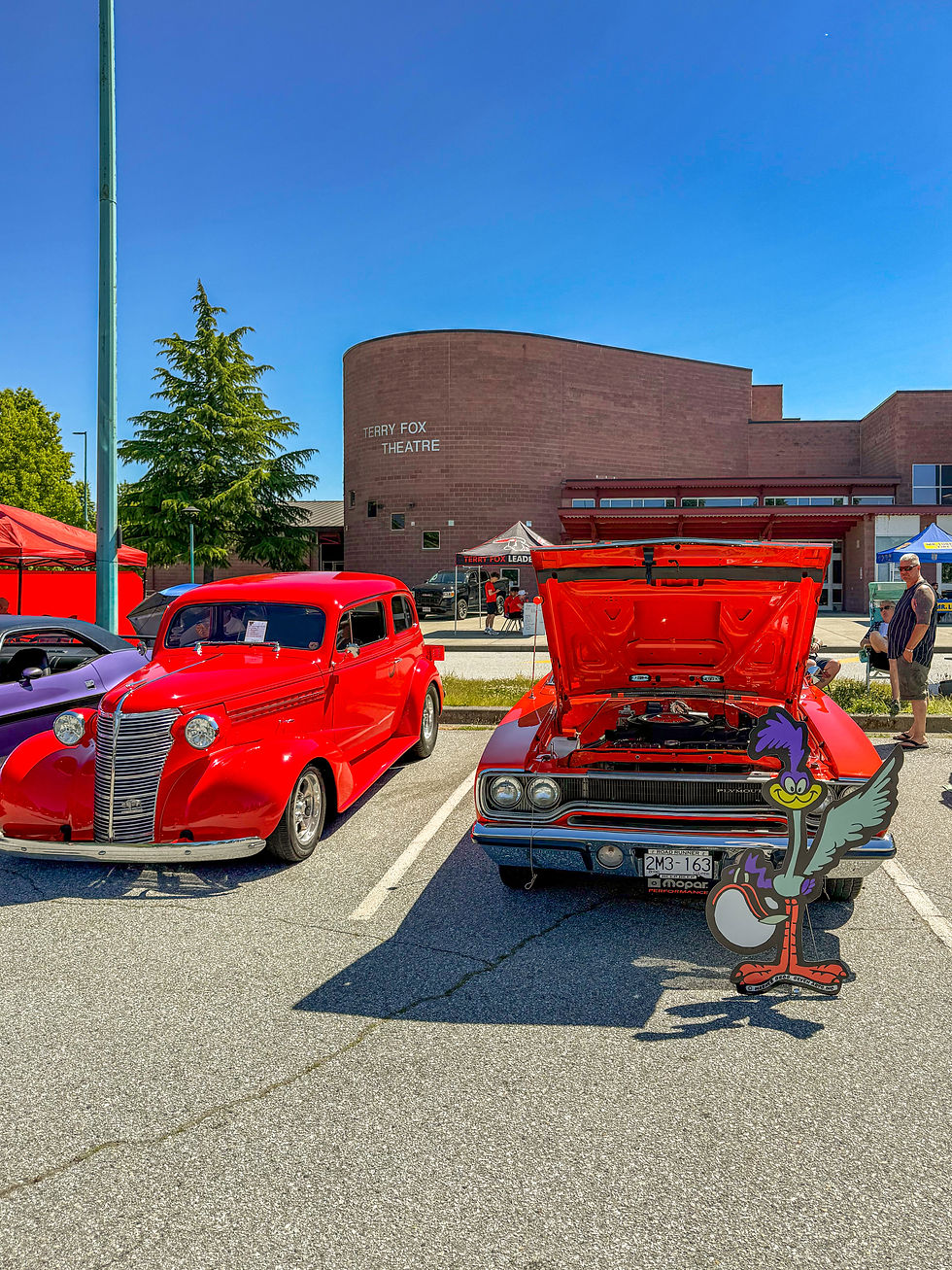 Classic red cars are displayed in a parking lot with the Terry Fox Theatre in the background. A Road Runner standee is in front of one car.