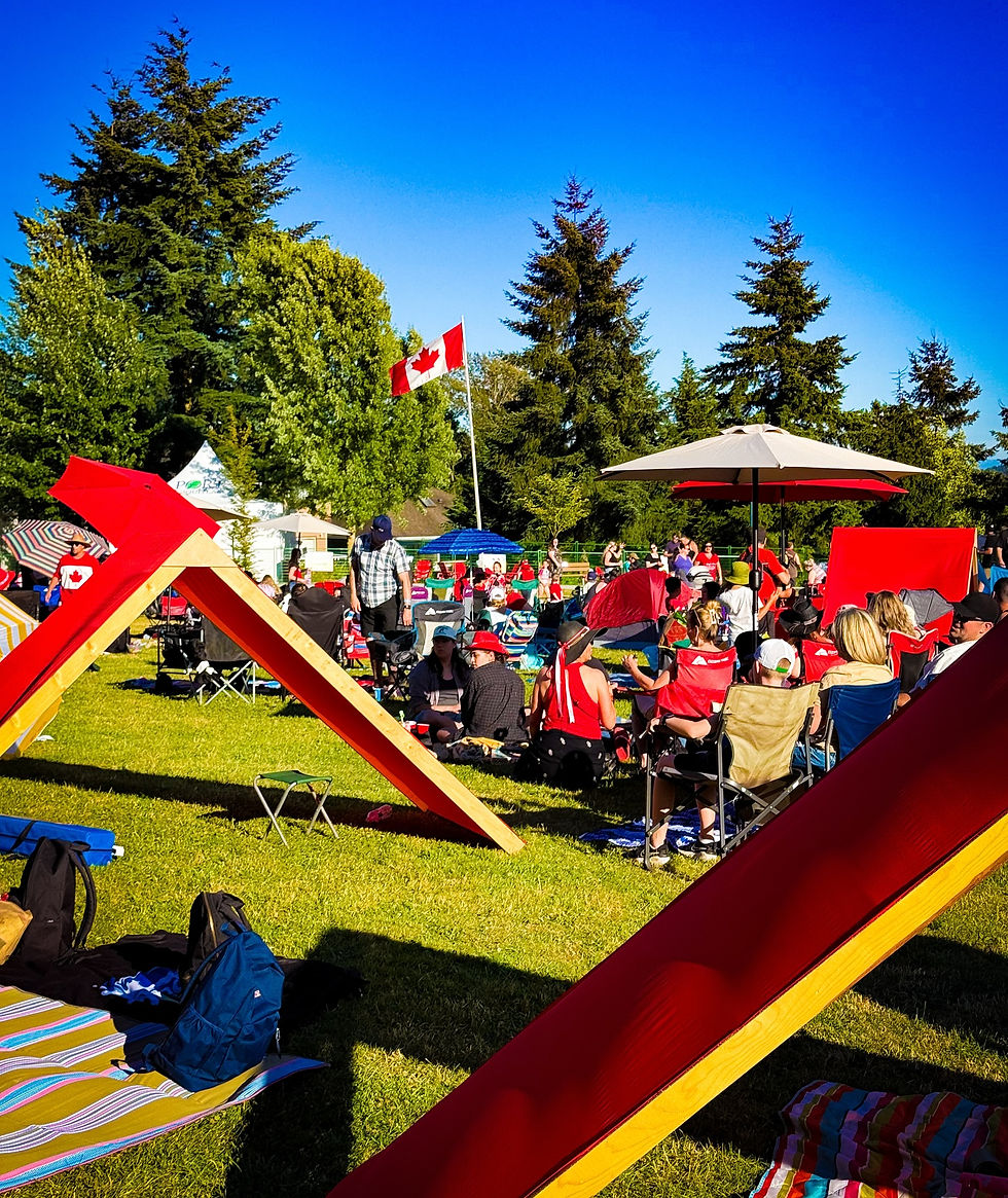 People in red clothing gather in a park under sunny skies with a Canadian flag in the background, surrounded by trees and picnic setups.