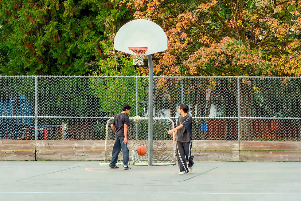 Two people playing basketball beneath a white backboard at Routley Park, with a small goal and autumn trees behind them.
