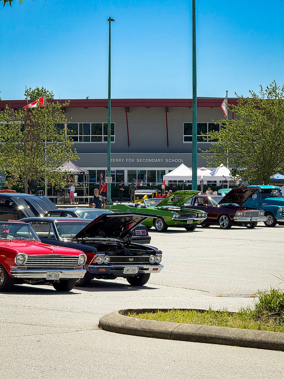 Classic cars with open hoods in a school parking lot. Terry Fox Secondary in the background. Bright day with flags visible.