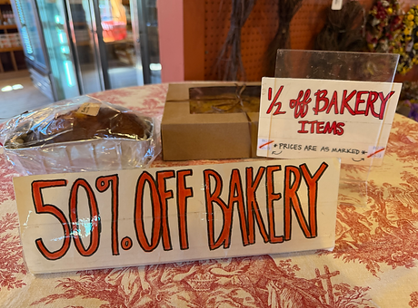 A table in the country store with a table cloth and 2 signs that say "50% OFF BAKERY" and "1/2 off BAKERY ITEMS *PRICES ARE AS MARKED* [barcode examples on both sides]" in red ink outlined in black on white paper