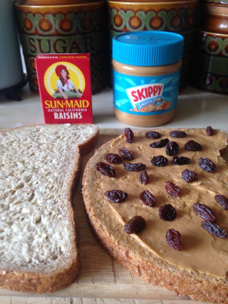 Sandwich being made on kitchen counter. Peanut butter spread with raisins on one slice, plain on the other. Skippy jar and raisins box nearby.