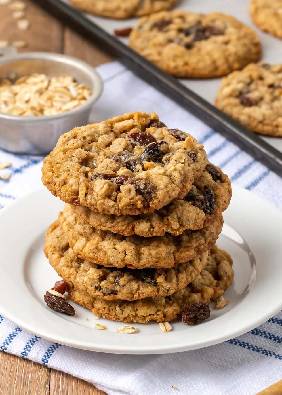 Stack of oatmeal raisin cookies on white plate, wooden table, striped cloth, bowl of oats, baking tray. Warm, homemade feel.