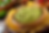 Bowl of green guacamole with red specks, surrounded by yellow tortilla chips on a wooden plate. Background shows blurred limes and tomatoes.