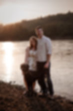 A tall white man with short dark hair has his arm around a short white woman with light brown shoulder length hair, a large curly haired dog stands by their feet. The background is the sunset rocky shore of a river. 