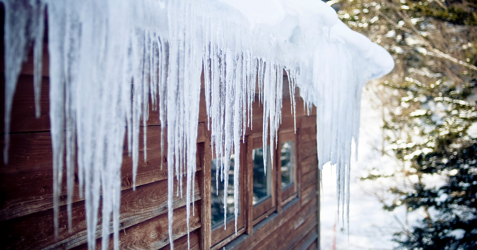 Icicles hang from a snowy roof of a wooden cabin. Snow-covered trees are in the background, creating a serene winter scene.