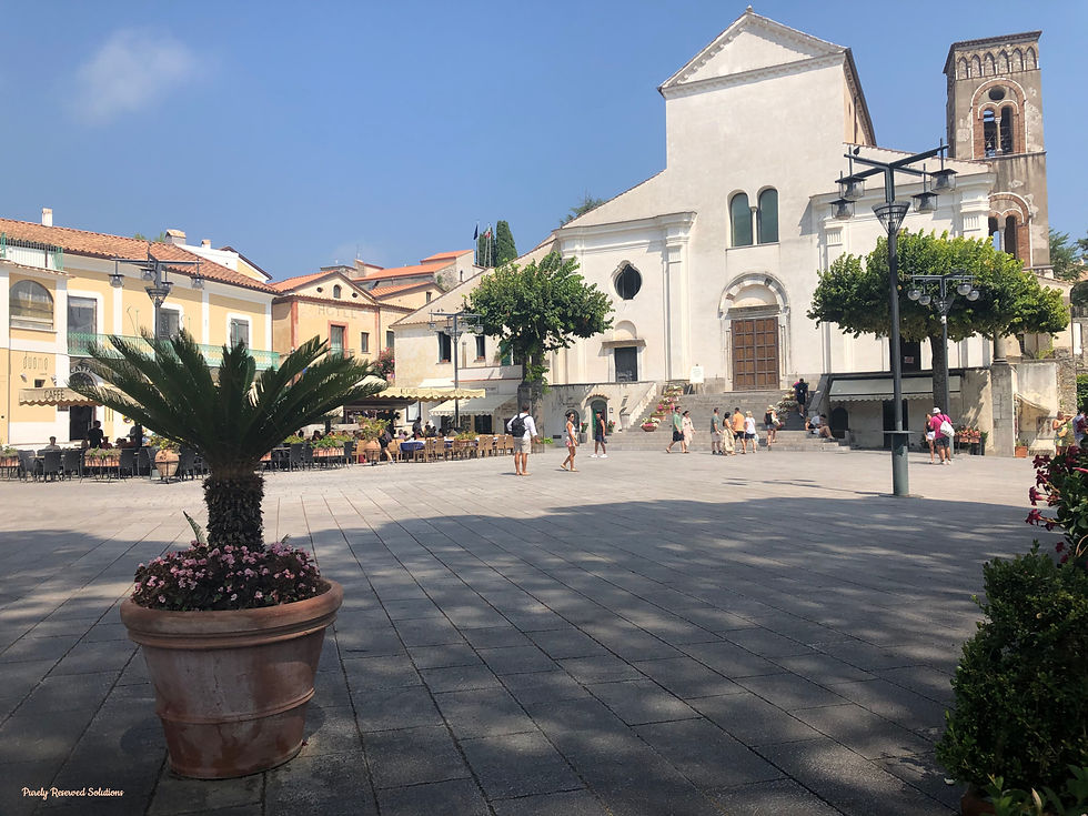 Piazza del Duomo in the heart of Ravello. Sunny town square with a potted palm in the foreground, people strolling near a white church, and cafés along the sides. Relaxed atmosphere.