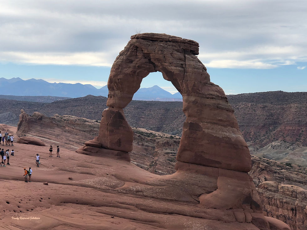 People explore near Delicate Arch, a large, natural sandstone arch in a rocky desert landscape. Mountains and cloudy skies are in the background.