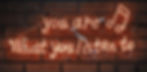Neon sign on brick wall reading "You are What you listen to" with musical note. Warm orange glow, urban setting, motivational mood.