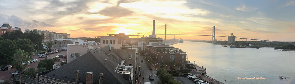 Sunset over a riverside cityscape with a prominent bridge, buildings, and parked cars. Warm hues light the sky, creating a calm mood.