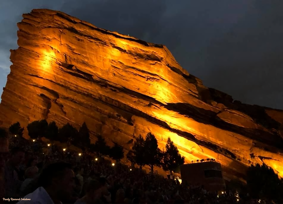 Illuminated rock formation against a dark sky, with a crowd below at the outdoor venue of Red Rocks Amphitheater. Warm lighting highlights the textured surface.