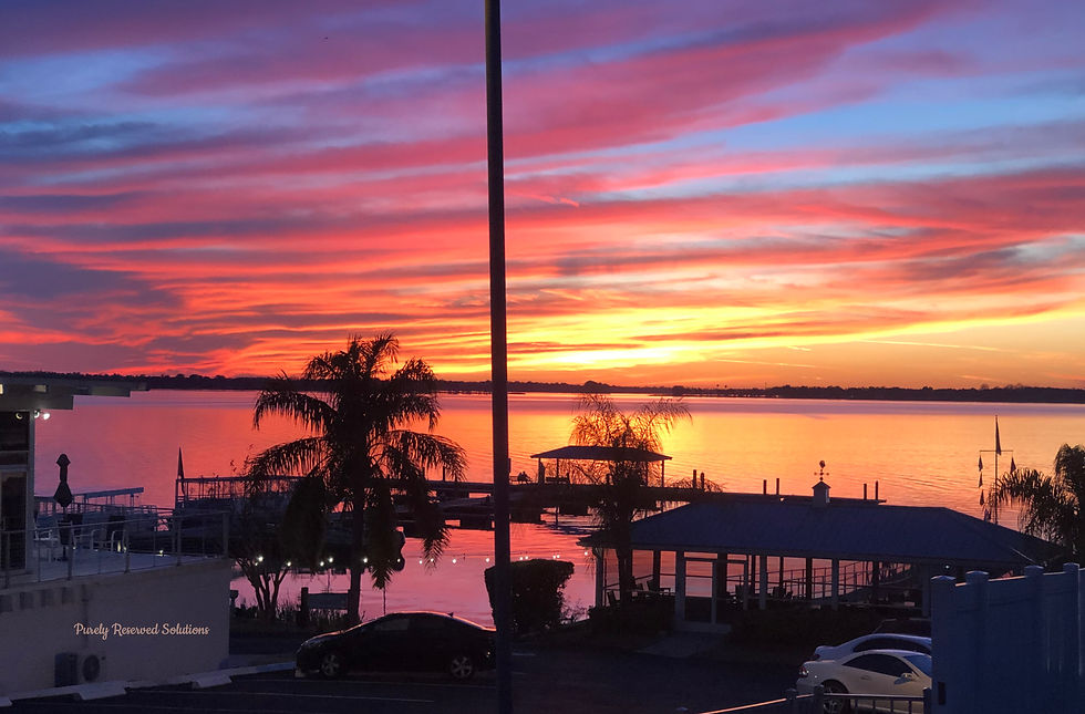 Vibrant sunset over a lake with silhouetted palm trees and docks. Orange, pink, and purple clouds fill the sky, creating a serene mood.