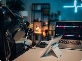 Microphone, laptop showing colorful audio waves, and tablet on desk. Cozy studio with shelves, candles, and neon blue line art in background.