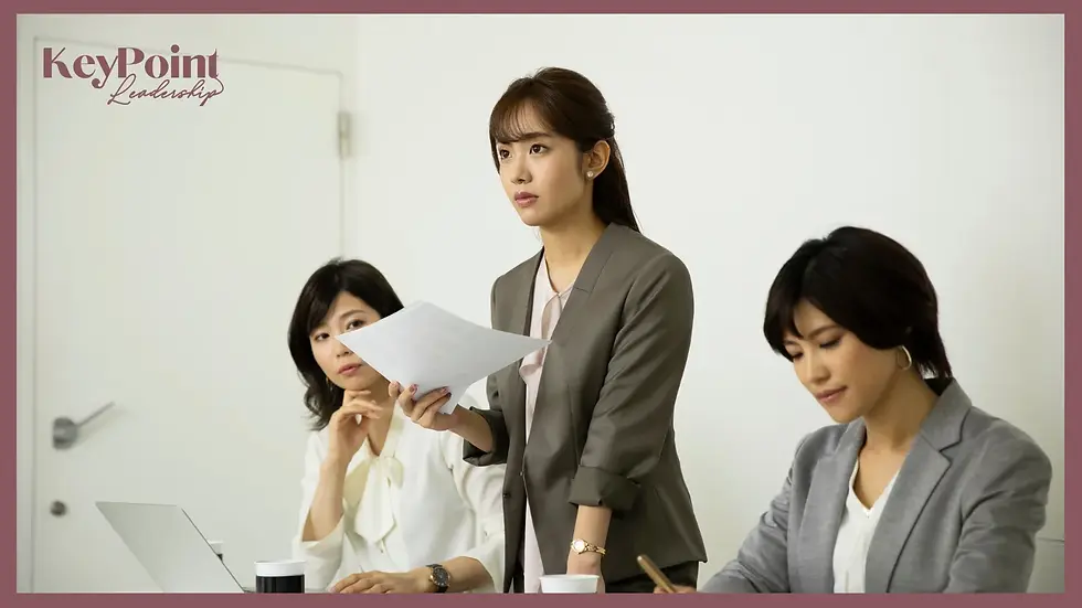 Three women in a meeting room; one stands holding papers, two sit with laptops. Neutral expressions, white wall background. Text: KeyPoint Leadership.