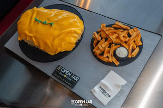 Oversized novelty parmo cake covered in melted cheese with fries and dip displayed at The Open Jar restaurant.