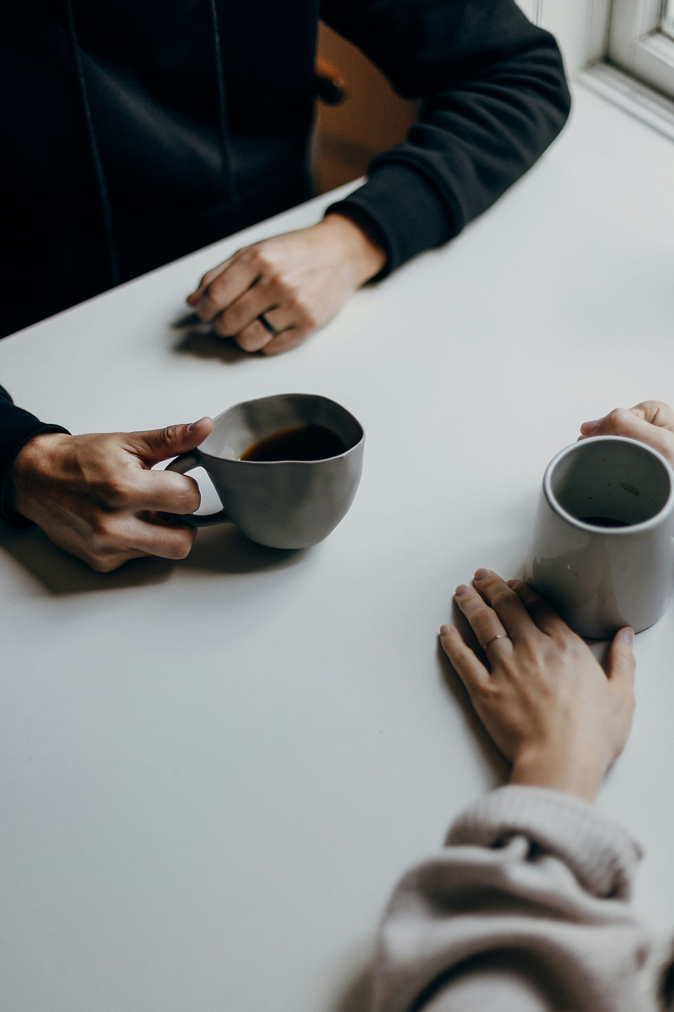 Two people’s hands around a white table holding coffee mugs, suggesting a quiet, casual conversation indoors.