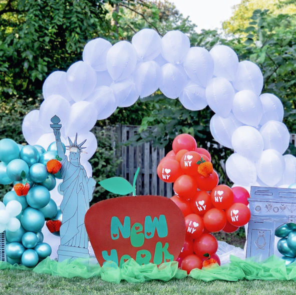 White balloon arch with red and teal balloon clusters, Statue of Liberty cutout, skyscraper props, and a big apple sign for a New York City–themed party decoration.