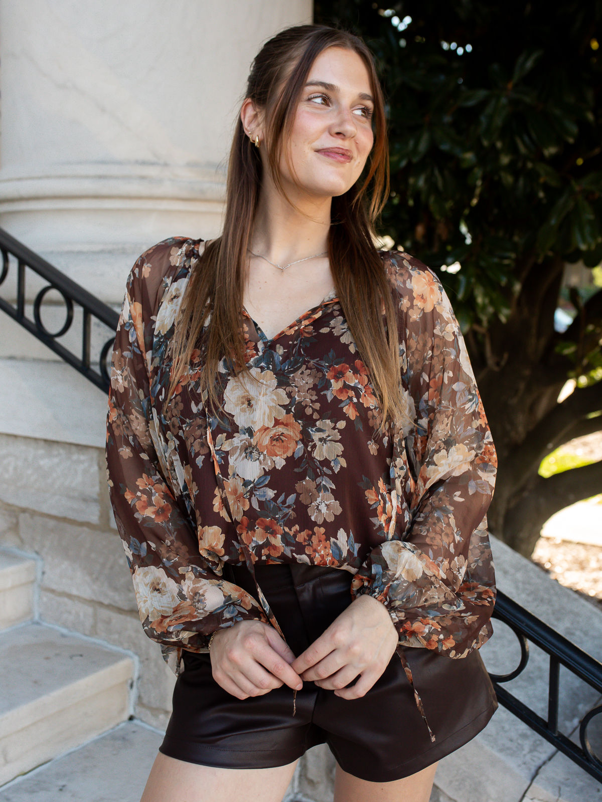 Model wearing the Camille Floral Blouse in Brown with rust, cream, and blue floral print, styled with dark faux leather short
