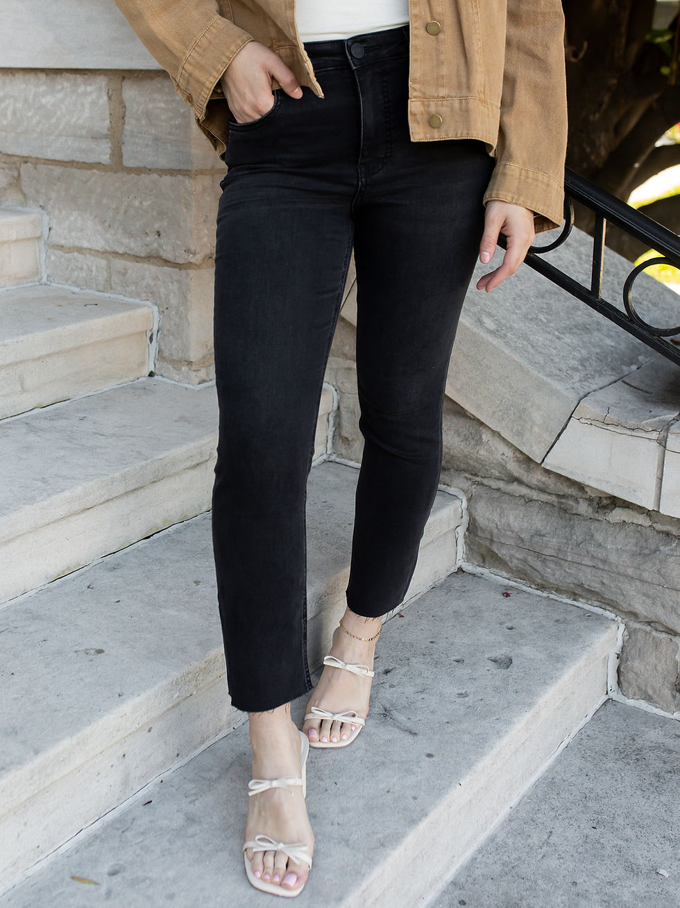 Model wearing Rachael High Rise Mom Jeans in Cleverly, featuring washed black denim, cropped raw hems, and a tan jacket.