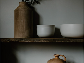 Japandi open shelving with rustic reclaimed wood, carved timber vase with green foliage, matte white ceramic bowls, and a clay terracotta jug.