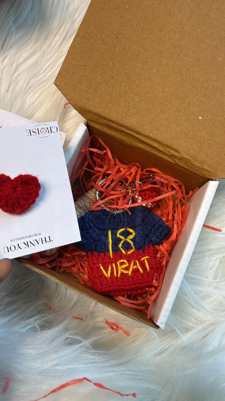 Open box with red shredded paper, blue-red knitted item labeled "18 VIRAT." White card with a red heart reads "Thank You." White furry background.