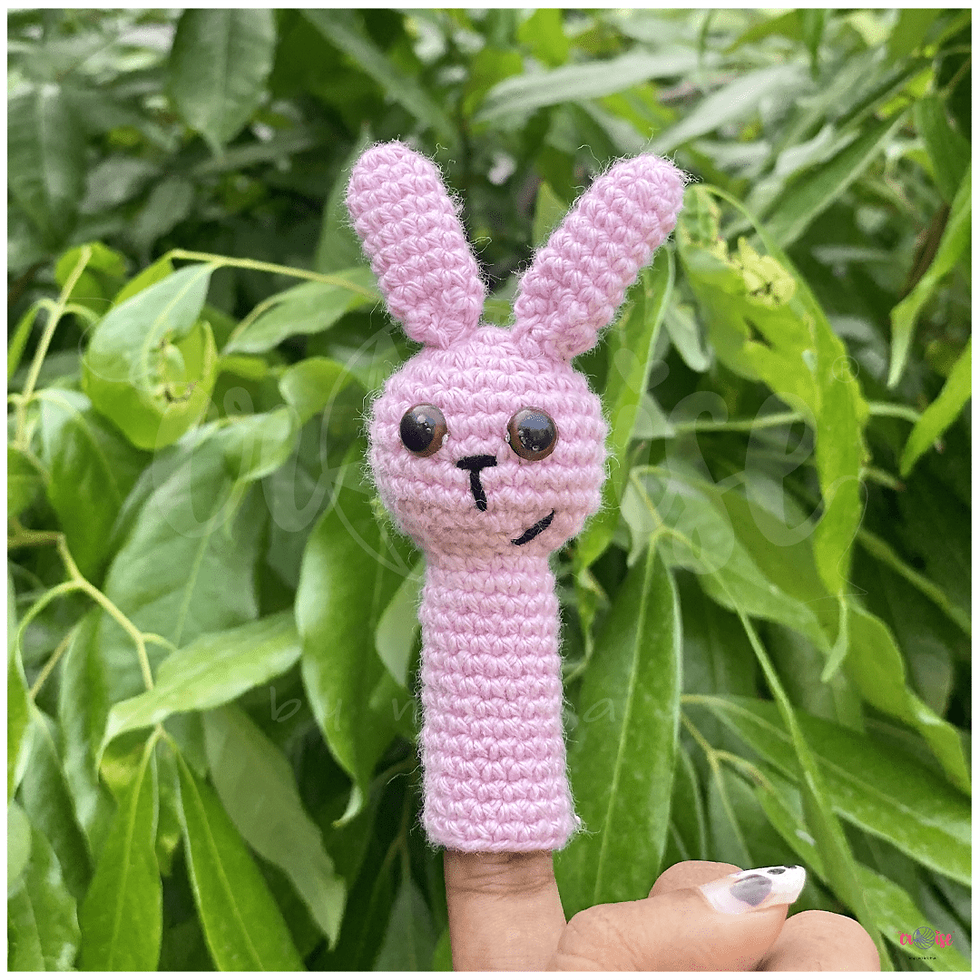 Crocheted gray donkey finger puppet with big eyes on a finger over a grassy background.