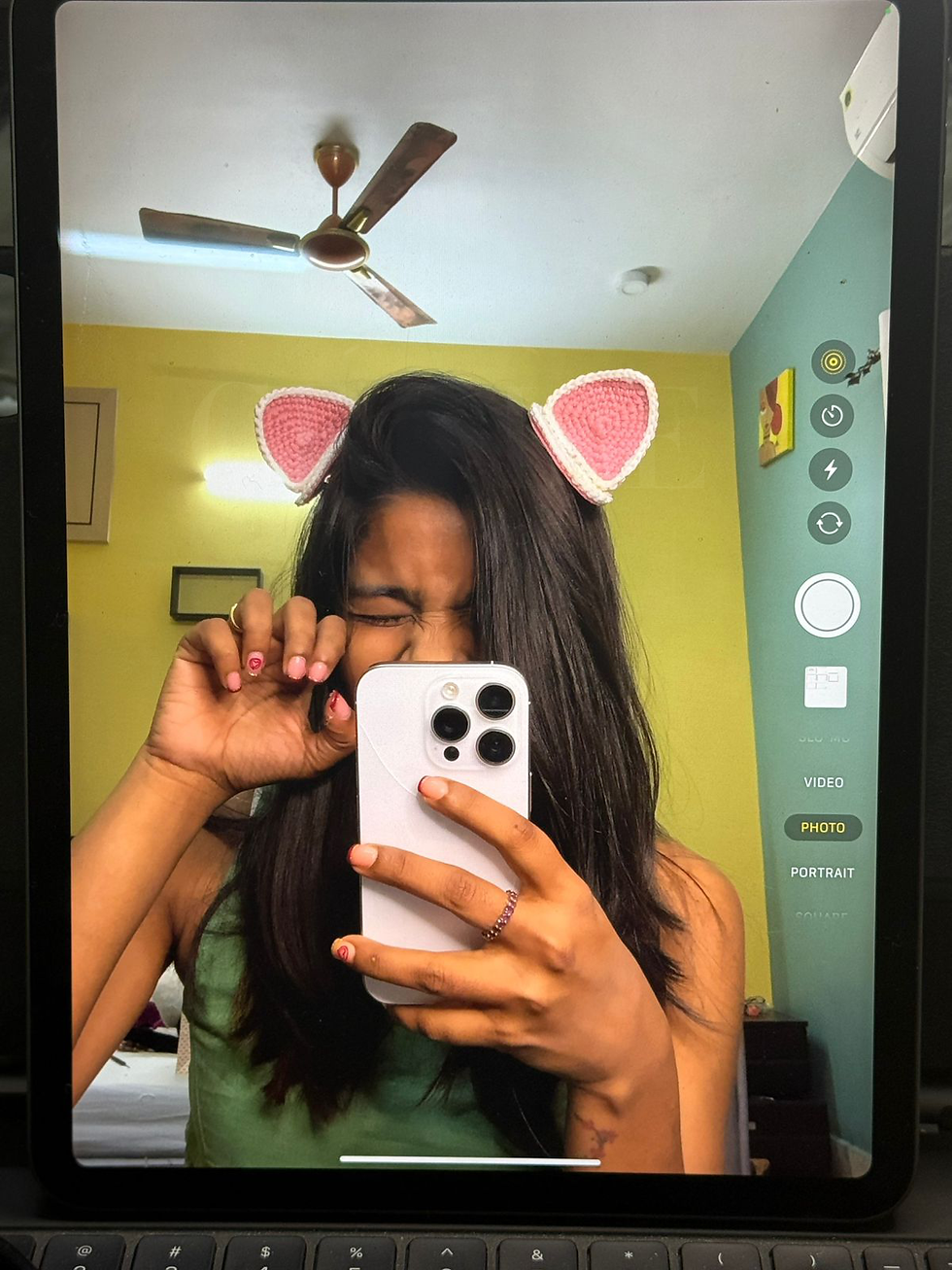 A woman wearing cat ear headbands takes a playful mirror selfie, winking and holding a phone. Background shows a colorful room with a ceiling fan.