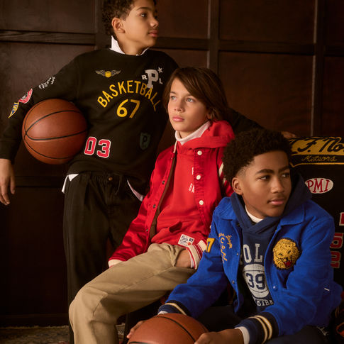 A group of 3 boys dressed in sporty and streetwear Polo Ralph Lauren. 2 Boys are sitting on a chair and one is standing all looking off camera.