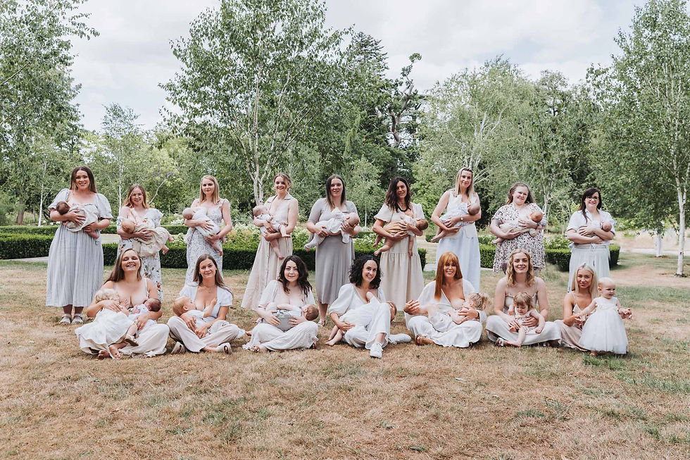 Women in light dresses sit and stand holding babies in a park. Green trees and grass create a serene backdrop, evoking a joyful mood.