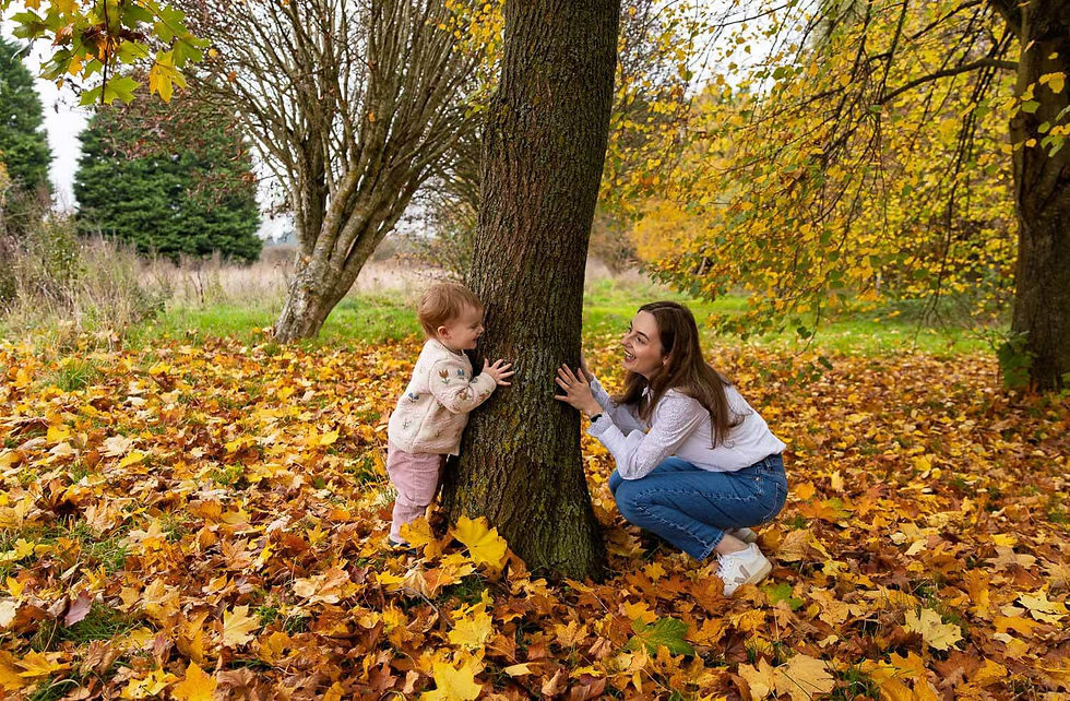 Woman and toddler play hide-and-seek around a tree in a park during autumn photoshoot in Worcester. Autumn leaves cover the ground. Both are smiling, surrounded by autumn foliage.