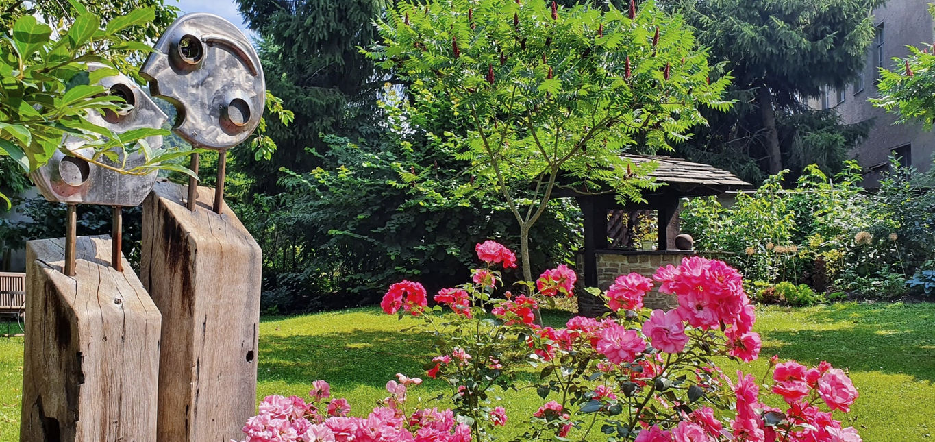 Panoramablick des Gartens mit Skulptur und blühenden Rosen im Vordergrund.
