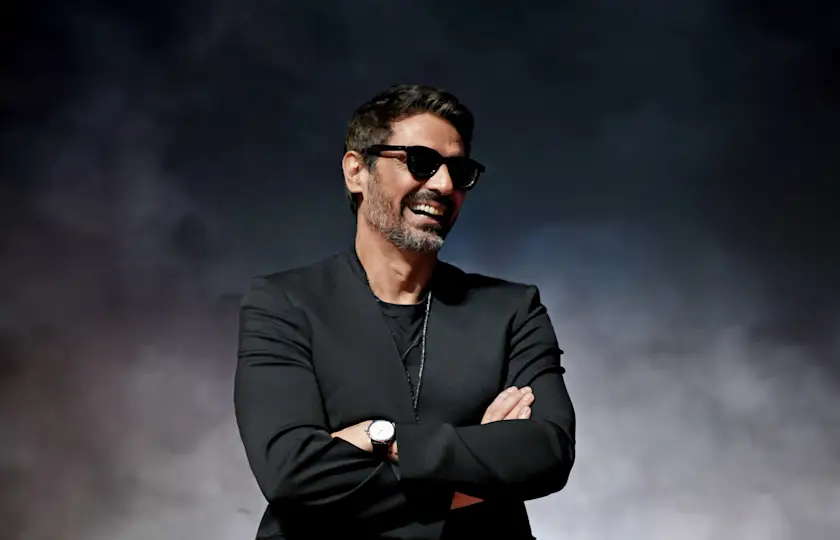 Smiling man in sunglasses and black jacket stands with arms crossed against a dark smoky background.