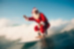 Santa Claus surfs a wave in a red suit under a clear blue sky. The ocean splashes around him, creating a festive, adventurous mood.