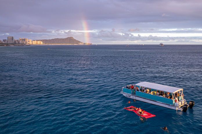 Golden sky reflected in calm seas as a boat sails with Diamond Head and a rainbow in the background.
