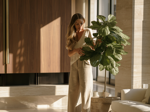 Woman arranging fake plants and artificial flowers in a modern dining room with warm wood, neutral decor, and natural light.