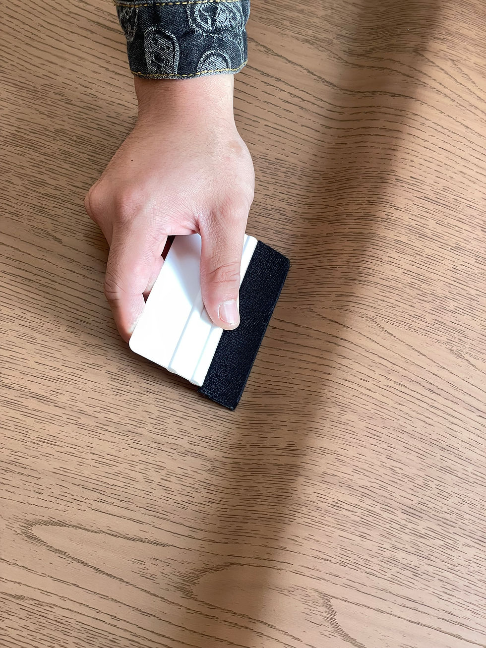 Hand in a patterned denim sleeve holding a white and black squeegee on a wood-grain table, casting a shadow.