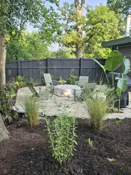 Stone patio with fire pit and chairs in a lush backyard.